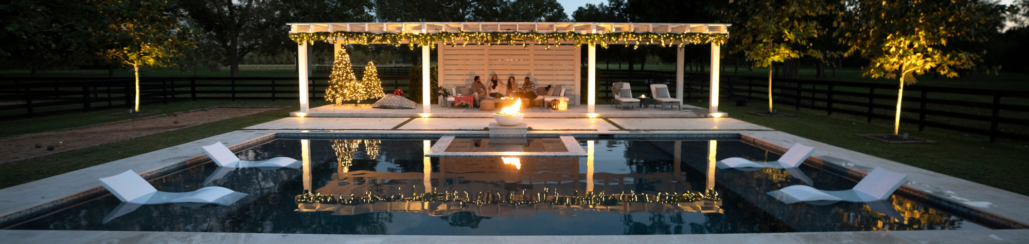 Four people relax on white lounge chairs in a modern swimming pool, surrounded by blue water, with patio furniture and a fire pit nearby. The scene is viewed from above.