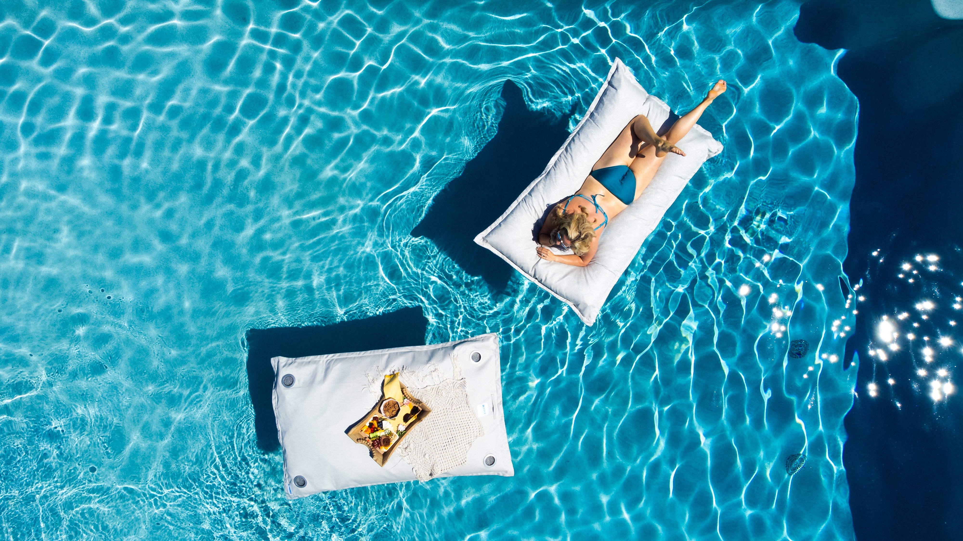 A woman in a color-blocked swimsuit relaxes on a blue float in a clear, turquoise pool, viewed from above.