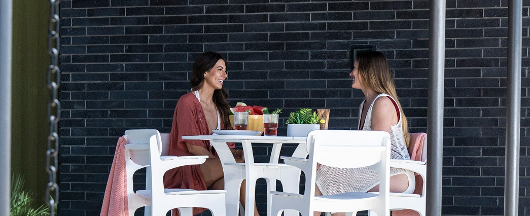 Two women sitting at a small outdoor dining table with white chairs, set against a dark brick wall background.