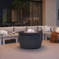 An outdoor patio scene features the Ledge Lounger Halo Firepit at its center, surrounded by a U-shaped sectional sofa with gray cushions. A book and blanket rest on the sofa, adding to the ambiance. Soft lighting and modern decor enhance this cozy setting.