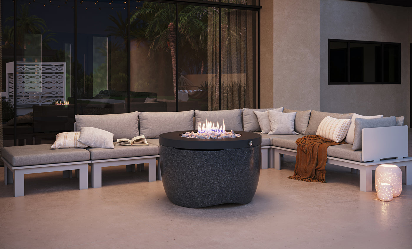 An outdoor patio scene features the Ledge Lounger Halo Firepit at its center, surrounded by a U-shaped sectional sofa with gray cushions. A book and blanket rest on the sofa, adding to the ambiance. Soft lighting and modern decor enhance this cozy setting.