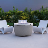 Three white, curved chairs are arranged around the Halo Firepit by Ledge Lounger on a light stone patio. The fire pit features a pivot-glide lid and emits a small flame, while lush green plants and soft ground lights decorate the background.