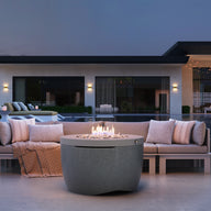 An outdoor seating area includes the sleek Halo Firepit by Ledge Lounger, surrounded by a beige sofa and cushions. Overlooking a pool and modern house with large windows under the twilight sky, green plants enhance the ambiance.
