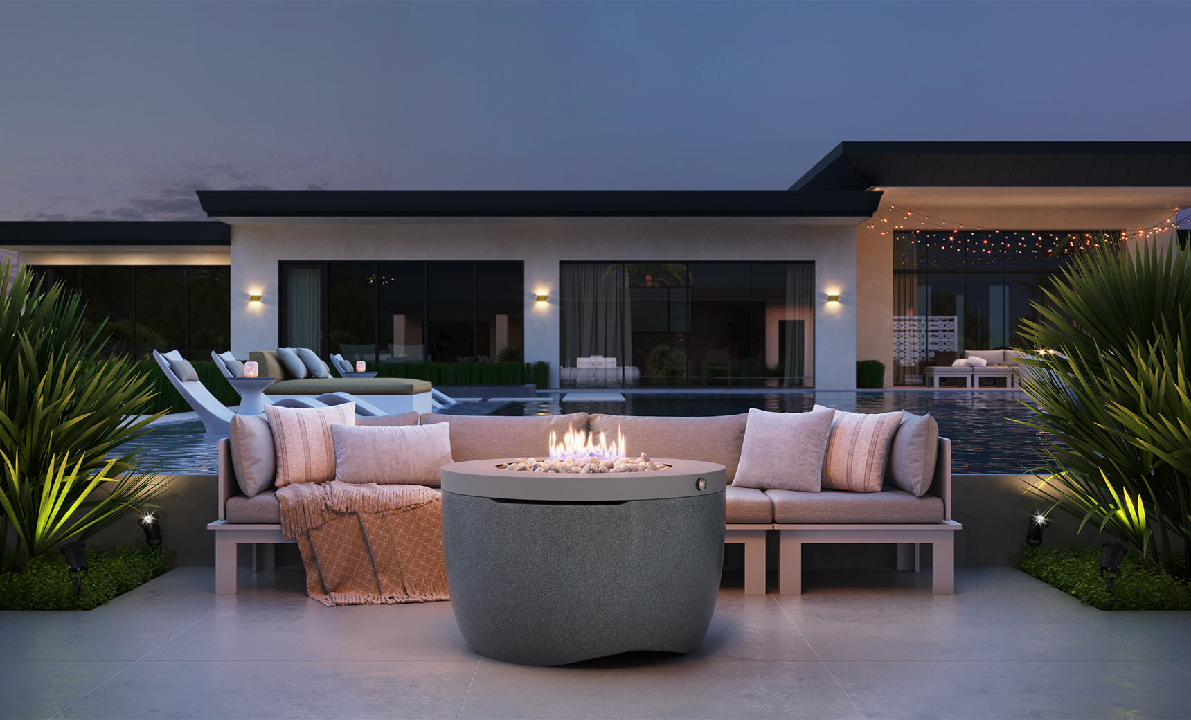 An outdoor seating area includes the sleek Halo Firepit by Ledge Lounger, surrounded by a beige sofa and cushions. Overlooking a pool and modern house with large windows under the twilight sky, green plants enhance the ambiance.
