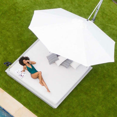 A woman in a teal swimsuit relaxes on a large white outdoor daybed under a large white umbrella. She is on her back, holding a magazine. The bed is on a grassy area next to a swimming pool.