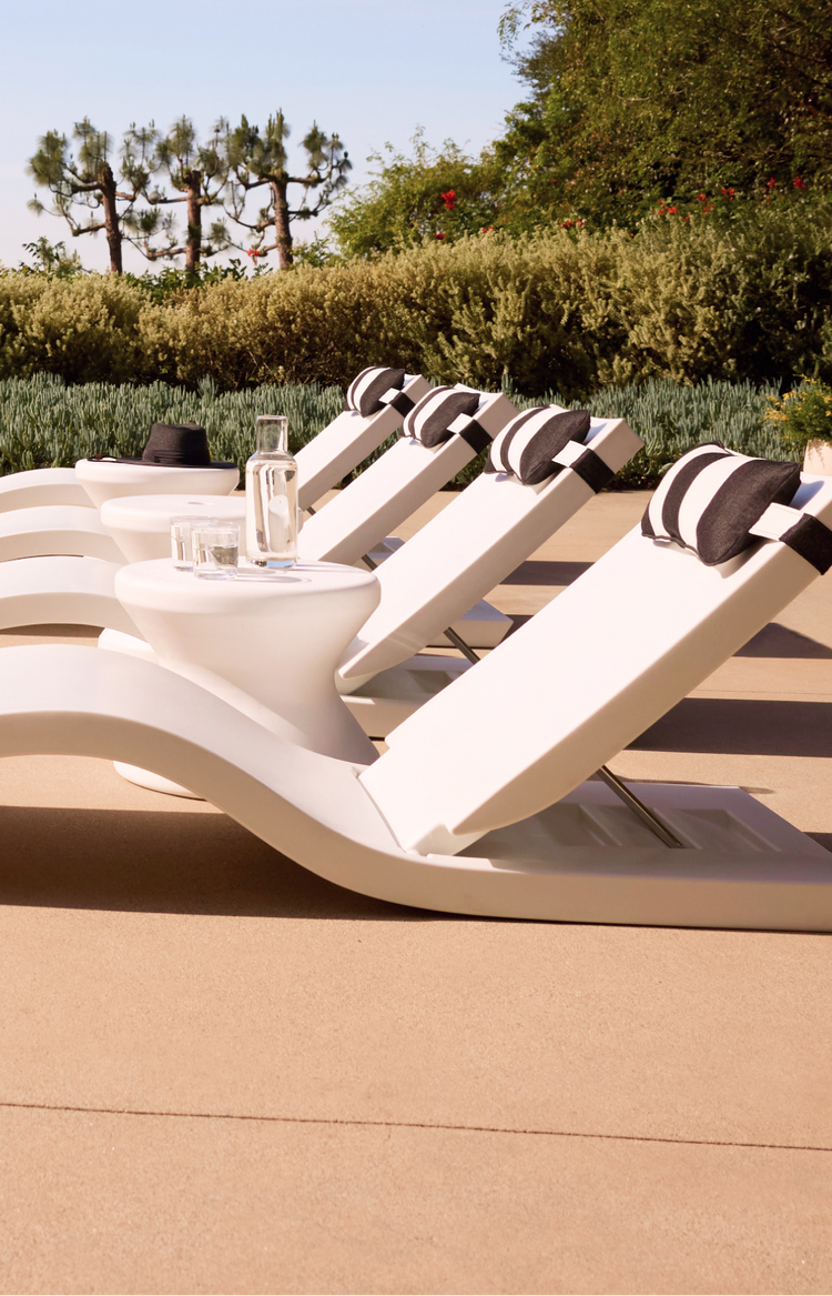White lounge chairs with black cushions on a sandy beach with trees in the background