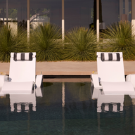 Two Ledge Lounger Upgrade Your Signature Chaise Armrest Consoles sit partially submerged in a pool, paired with black-and-white pillows, offering patio comfort among tall grasses and a backdrop of glass windows.