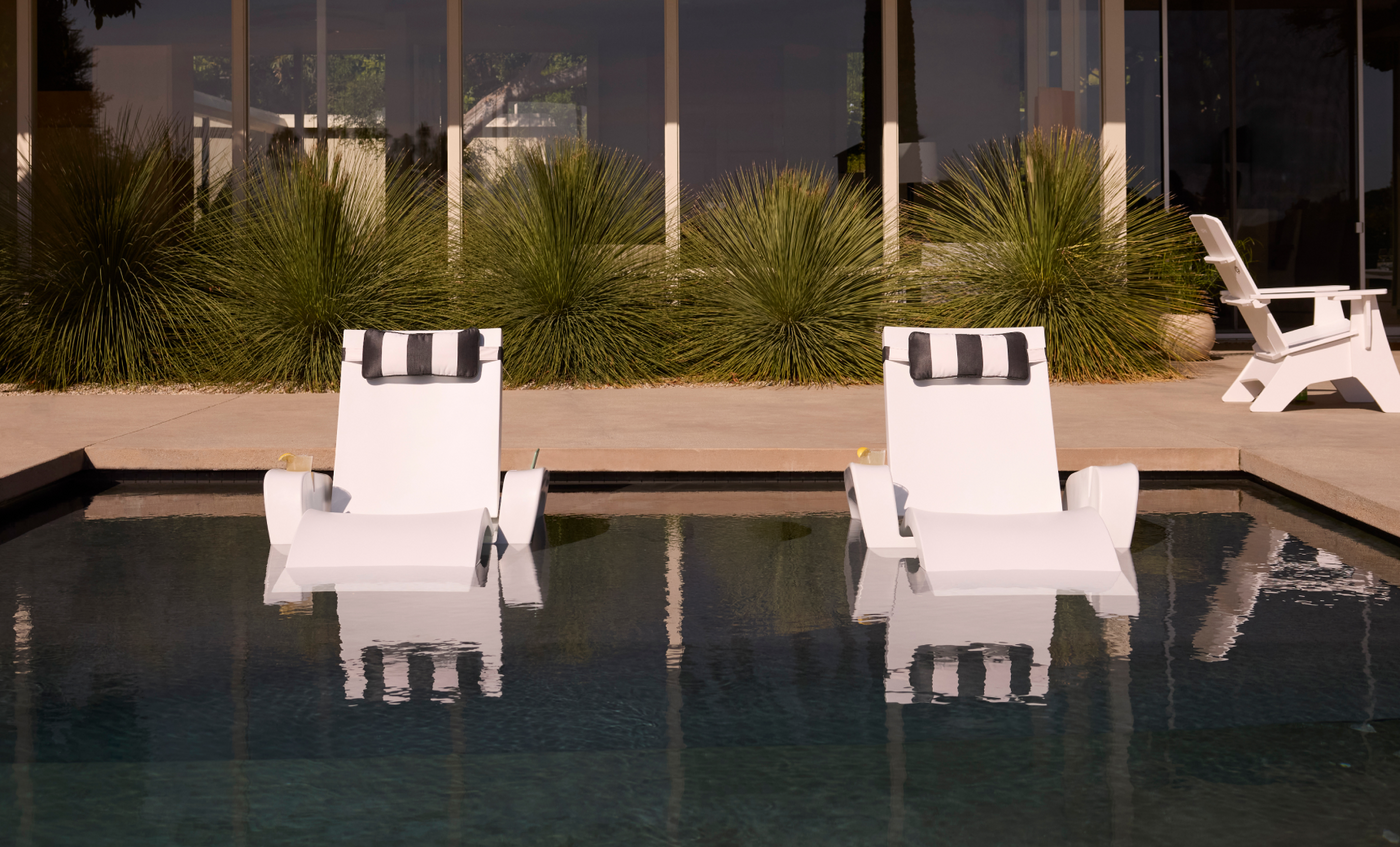 Two Ledge Lounger Upgrade Your Signature Chaise Armrest Consoles sit partially submerged in a pool, paired with black-and-white pillows, offering patio comfort among tall grasses and a backdrop of glass windows.