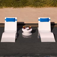 The Ledge Lounger Adjustable Chaise with Pillows and Side Table In-Pool & Patio Set features two white chaises with blue pillows and a matching table, perfect for relaxing in a shallow pool. Ornamental grasses and a concrete walkway complete the serene backdrop.