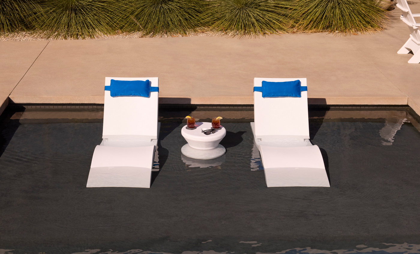 The Ledge Lounger Adjustable Chaise with Pillows and Side Table In-Pool & Patio Set features two white chaises with blue pillows and a matching table, perfect for relaxing in a shallow pool. Ornamental grasses and a concrete walkway complete the serene backdrop.
