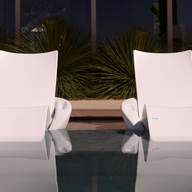 Two Ledge Lounger Upgrade Your Signature Chaise Armrest Consoles (2pk) sit side by side, partially submerged in the pool. Greenery and glass windows frame the tranquil scene, with reflections adding to the serene, patio-style comfort.
