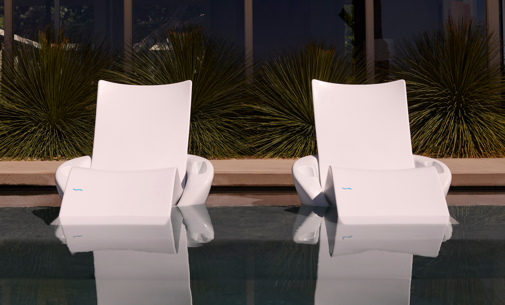 Two Ledge Lounger Upgrade Your Signature Chaise Armrest Consoles (2pk) sit side by side, partially submerged in the pool. Greenery and glass windows frame the tranquil scene, with reflections adding to the serene, patio-style comfort.