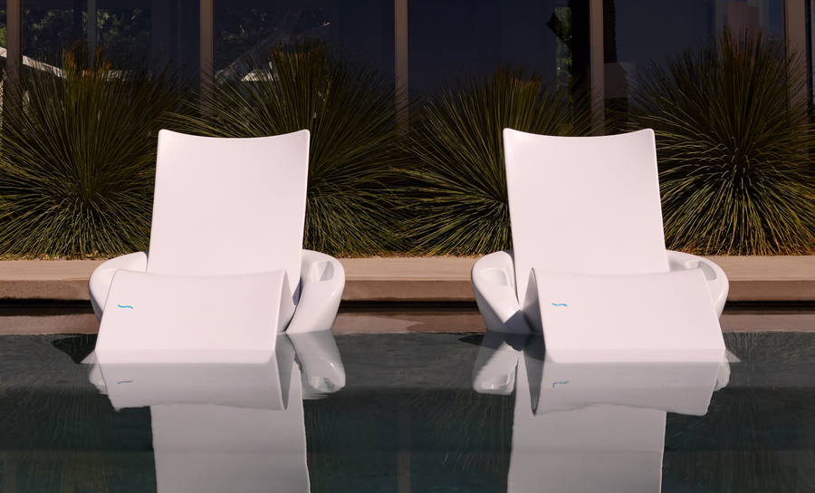 Two Ledge Lounger Upgrade Your Signature Chaise Armrest Consoles (2pk) sit side by side, partially submerged in the pool. Greenery and glass windows frame the tranquil scene, with reflections adding to the serene, patio-style comfort.