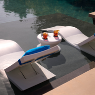 The Ledge Lounger Adjustable Chaise with Pillows and Side Table In-Pool & Patio Set features two modern white loungers with blue cushions, partially submerged poolside beside a round table—ideal for stylish outdoor relaxation.