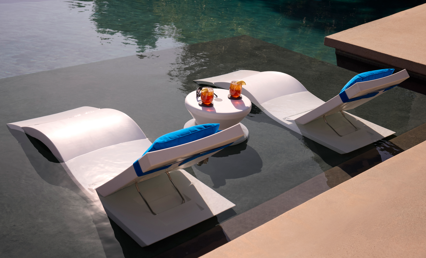 The Ledge Lounger Adjustable Chaise with Pillows and Side Table In-Pool & Patio Set features two modern white loungers with blue cushions, partially submerged poolside beside a round table—ideal for stylish outdoor relaxation.
