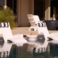 New description: The Ledge Lounger Signature Chaise Armrest Console features two white lounge chairs with black-and-white pillows in a pool beside a round table, with more white chairs on a sunny patio near greenery for ultimate relaxation.