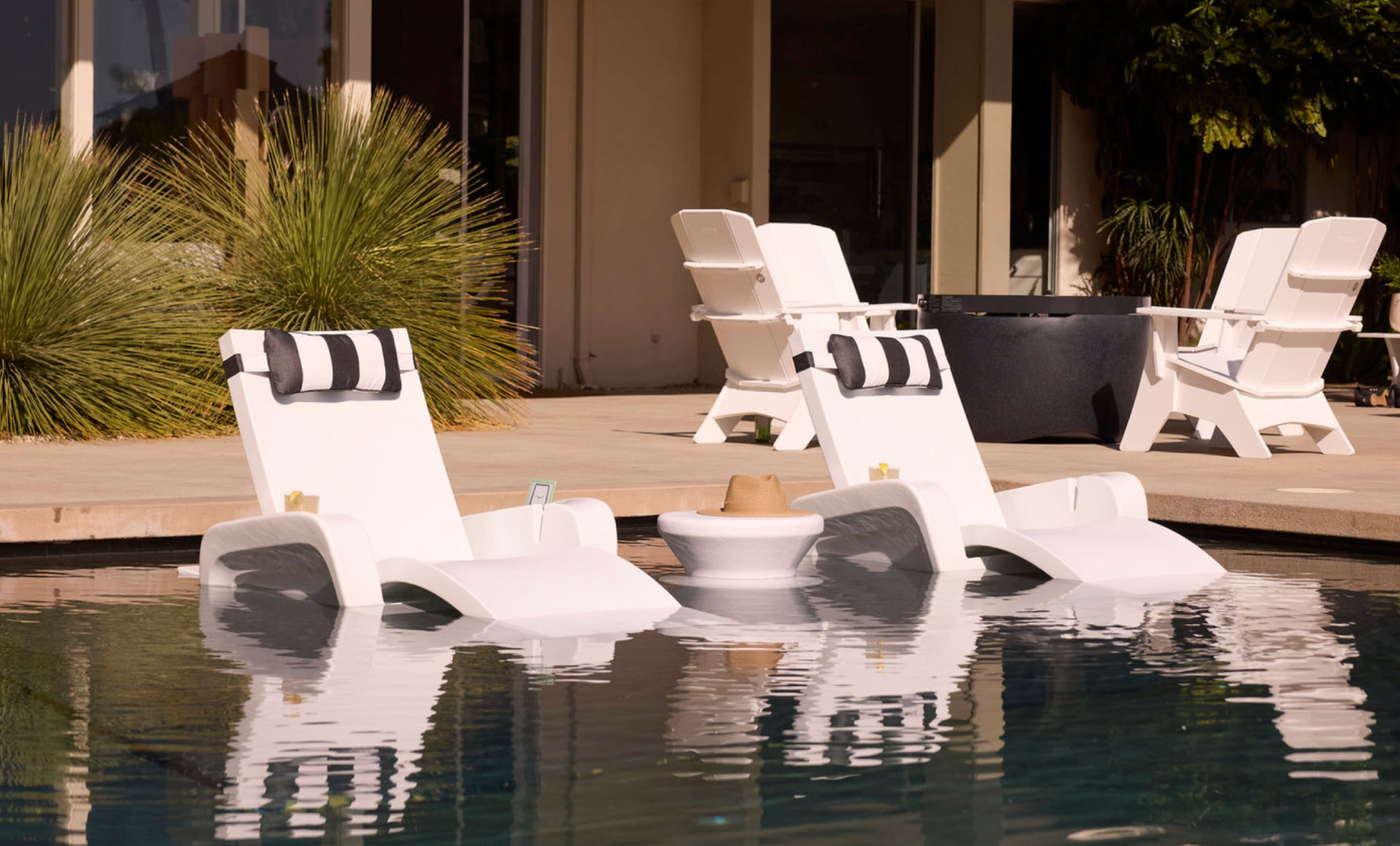 New description: The Ledge Lounger Signature Chaise Armrest Console features two white lounge chairs with black-and-white pillows in a pool beside a round table, with more white chairs on a sunny patio near greenery for ultimate relaxation.