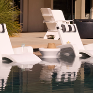 Two white Ledge Lounger Signature Chaises rest on a sun shelf in the pool, with a Signature Chaise Armrest Console between them holding a hat. More chaises are visible on the patio in the background.