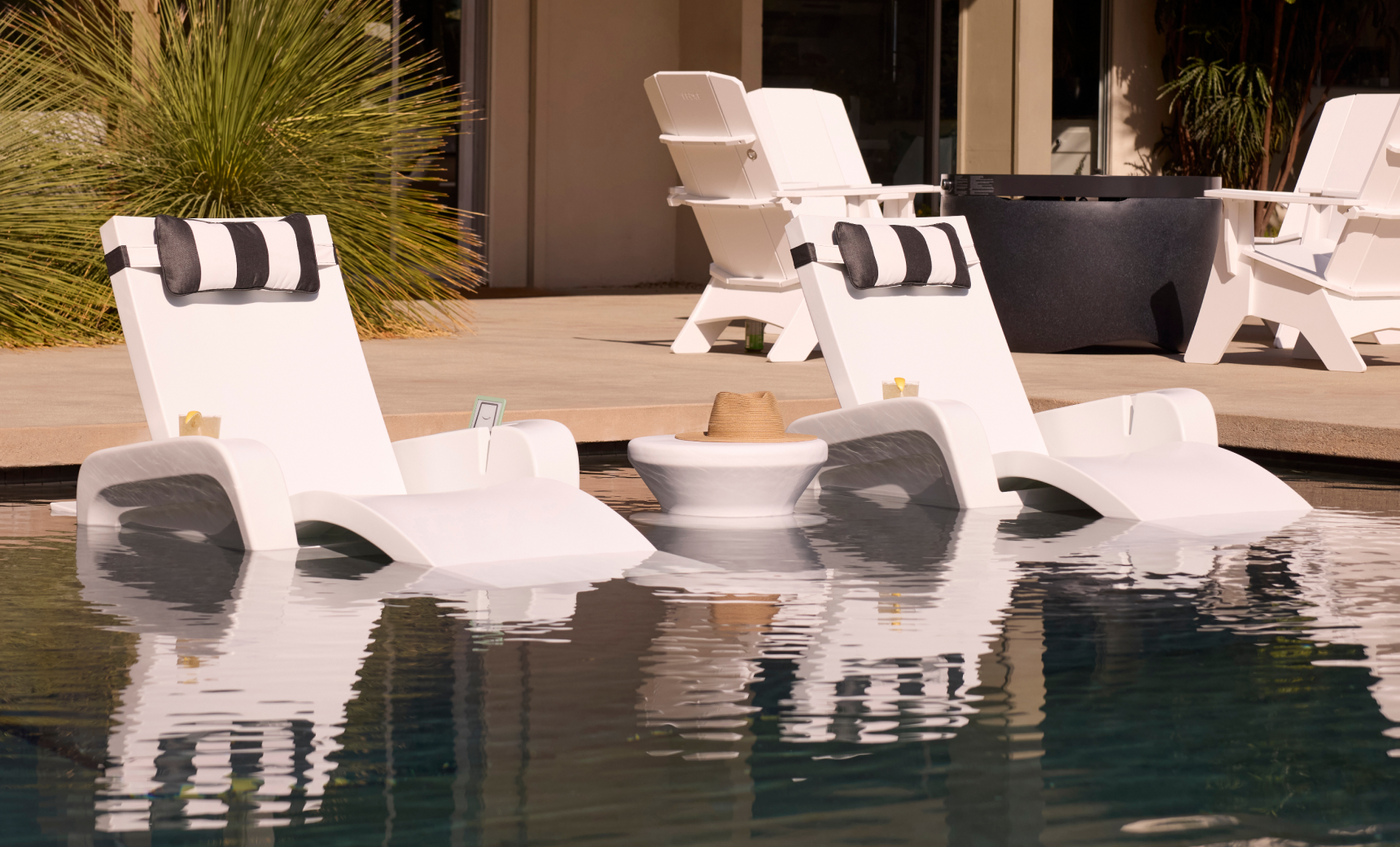 Two white Ledge Lounger Signature Chaises rest on a sun shelf in the pool, with a Signature Chaise Armrest Console between them holding a hat. More chaises are visible on the patio in the background.