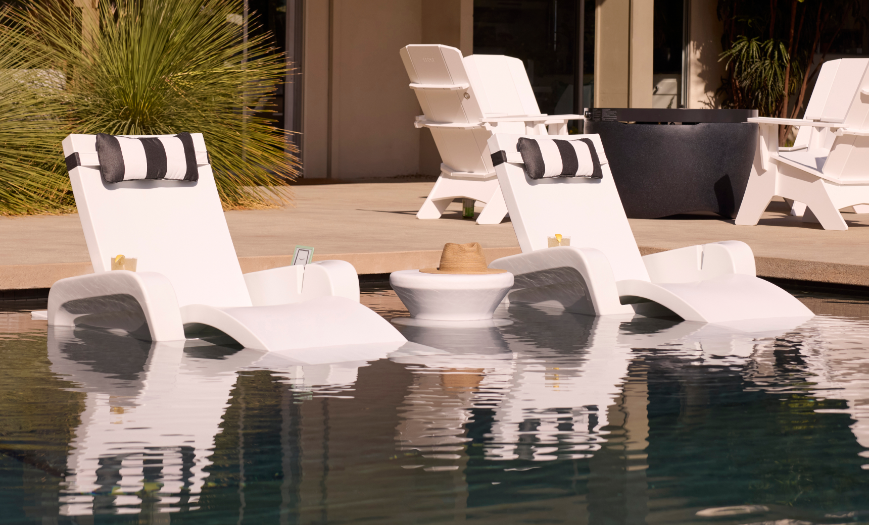 Two white Ledge Lounger Signature Chaises rest on a sun shelf in the pool, with a Signature Chaise Armrest Console between them holding a hat. More chaises are visible on the patio in the background.