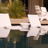 Three Ledge Lounger Signature Adjustable Reclining Chaises and a small round table sit partly submerged on the sun shelf, with patio chairs and green plants in the sunny background.