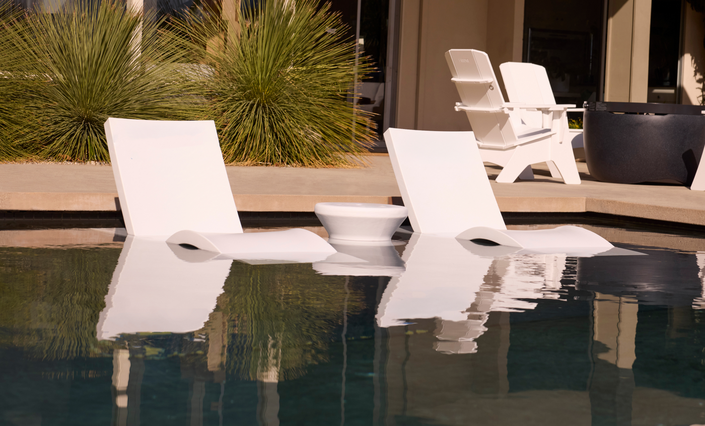 Three Ledge Lounger Signature Adjustable Reclining Chaises and a small round table sit partly submerged on the sun shelf, with patio chairs and green plants in the sunny background.