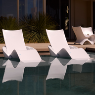 Two Ledge Lounger Autograph Chaise Armrest Consoles, in modern white, rest on a tanning ledge partially submerged in a pool. Reflections shimmer as tall green plants and more white chairs accent the scene near a contemporary building with glass doors.