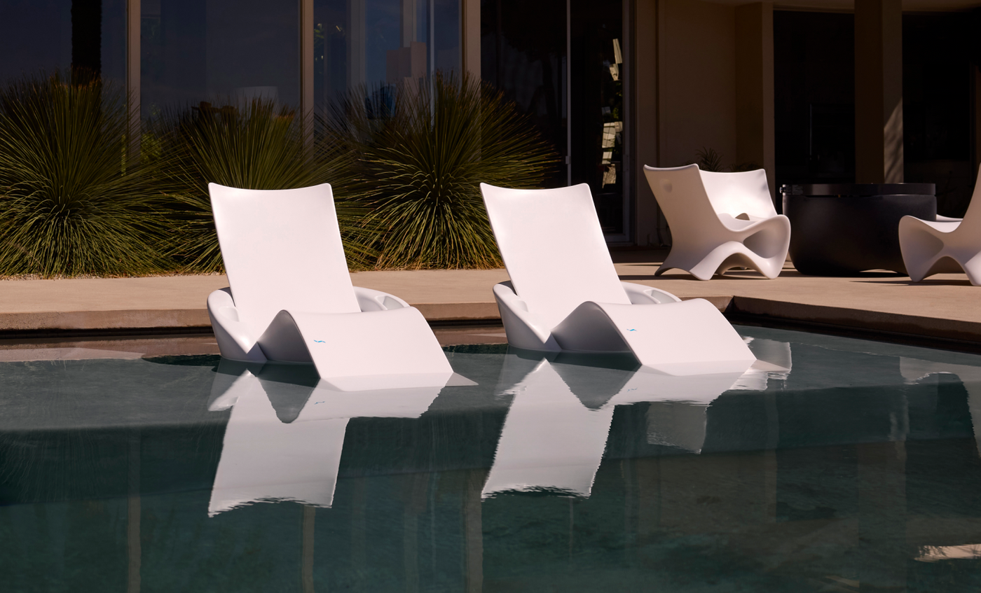 Two Ledge Lounger Autograph Chaise Armrest Consoles, in modern white, rest on a tanning ledge partially submerged in a pool. Reflections shimmer as tall green plants and more white chairs accent the scene near a contemporary building with glass doors.