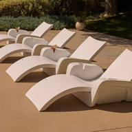 New description: Four Ledge Lounger Signature Chaise Armrest Consoles, modern white lounge chairs each with drinks on their armrests, are arranged beside a pool on a smooth tan patio amid greenery and new outdoor furniture.