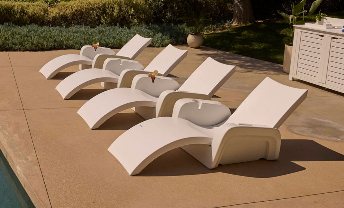 New description: Four Ledge Lounger Signature Chaise Armrest Consoles, modern white lounge chairs each with drinks on their armrests, are arranged beside a pool on a smooth tan patio amid greenery and new outdoor furniture.