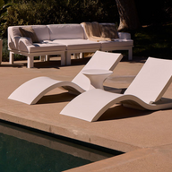 Two Signature Adjustable Reclining Chaises by Ledge Lounger in crisp white sit poolside on a concrete patio, ideal for relaxing after sunbathing, with a cushioned white outdoor sofa and potted plant visible in the background.