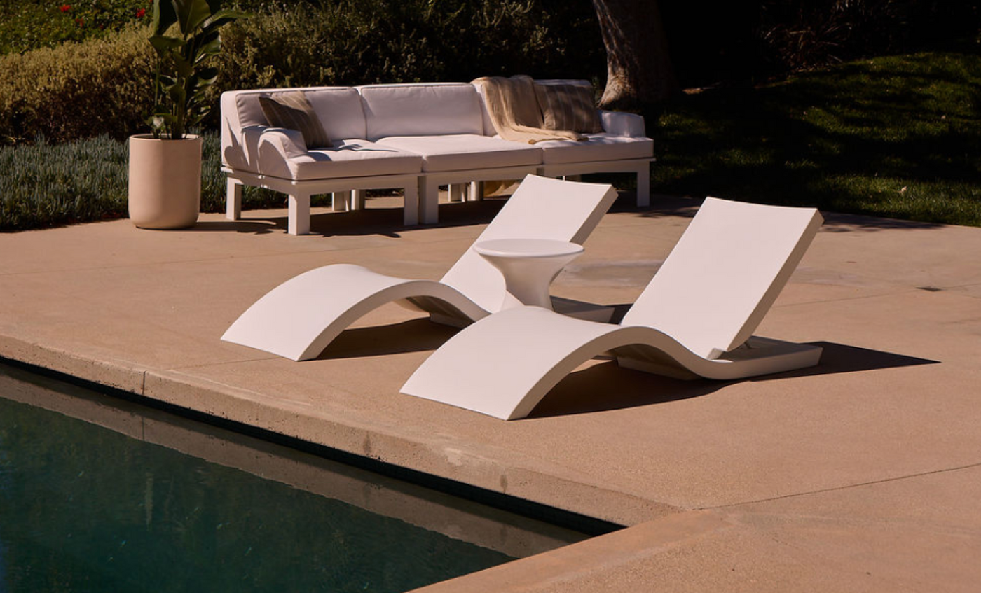 Two Signature Adjustable Reclining Chaises by Ledge Lounger in crisp white sit poolside on a concrete patio, ideal for relaxing after sunbathing, with a cushioned white outdoor sofa and potted plant visible in the background.