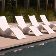 Four Ledge Lounger Autograph Chaise Armrest Consoles are lined up poolside on a concrete patio, ideal for relaxing near the tanning ledge with tall green plants and a modern building featuring large windows in the background.
