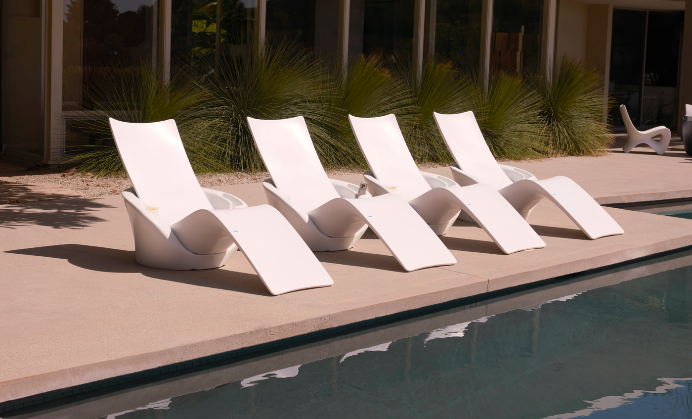 Four Ledge Lounger Autograph Chaise Armrest Consoles are lined up poolside on a concrete patio, ideal for relaxing near the tanning ledge with tall green plants and a modern building featuring large windows in the background.