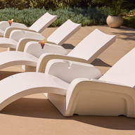 Four Ledge Lounger Signature Chaise Armrest Consoles are lined up on a sunlit patio, each holding a drink. Greenery and a large potted plant in the background create a relaxing vibe.