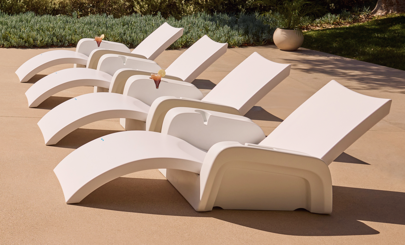 Four Ledge Lounger Signature Chaise Armrest Consoles are lined up on a sunlit patio, each holding a drink. Greenery and a large potted plant in the background create a relaxing vibe.