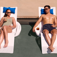 A woman and a man relax on Ledge Lounger’s Autograph Chaise Armrest Console on a pool's tanning ledge, both in sunglasses. The woman holds a drink, blue cushions rest under their heads, with the poolside area visible in the background.