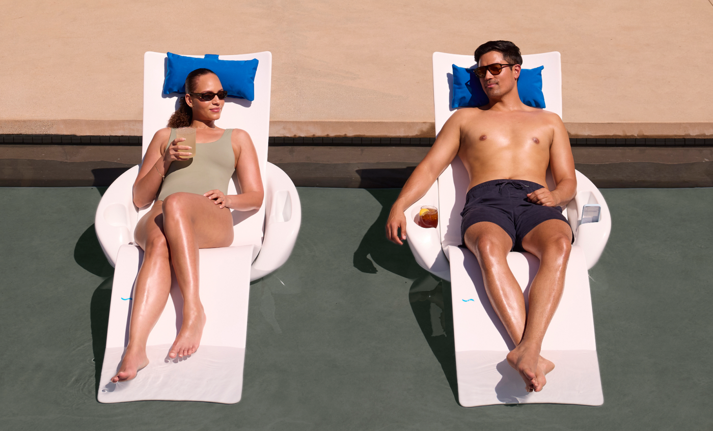 A woman and a man relax on Ledge Lounger’s Autograph Chaise Armrest Console on a pool's tanning ledge, both in sunglasses. The woman holds a drink, blue cushions rest under their heads, with the poolside area visible in the background.