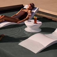 A woman in a swimsuit relaxes on a Ledge Lounger Signature Adjustable Reclining Chaise with a drink, while a man kneels on the sun shelf nearby. Next to them are an empty white chair and a small round table with two drinks.