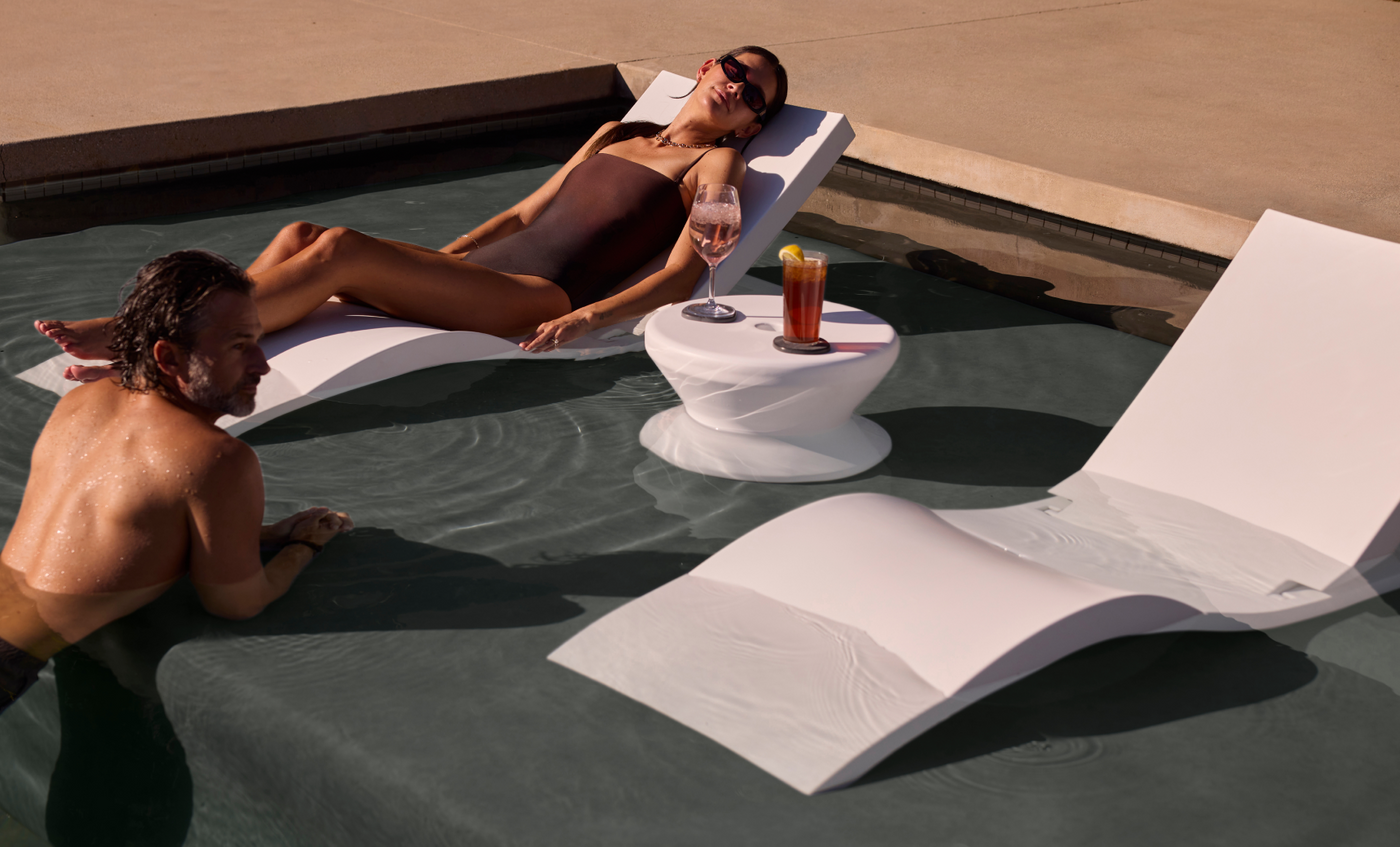 A woman in a swimsuit relaxes on a Ledge Lounger Signature Adjustable Reclining Chaise with a drink, while a man kneels on the sun shelf nearby. Next to them are an empty white chair and a small round table with two drinks.
