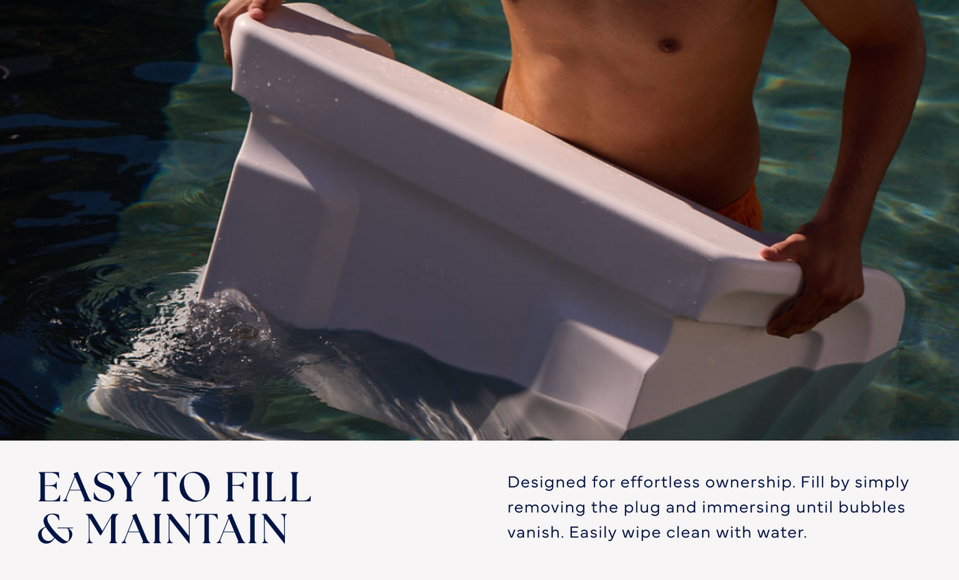 A person stands in a pool holding the Ledge Lounger Signature Chaise Armrest Console, partially submerged. It features easy filling and maintenance, with clear care instructions included for effortless use.