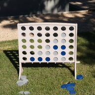 A Ledge Lounger Four In A Row oversized outdoor game stands on the grass, its all-weather board filled with blue and gray discs while a few pieces are scattered beneath in the sunlight.