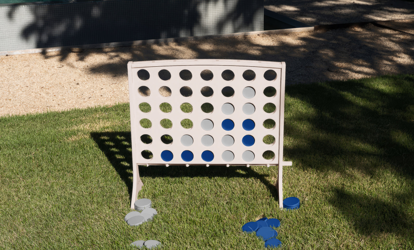 A Ledge Lounger Four In A Row oversized outdoor game stands on the grass, its all-weather board filled with blue and gray discs while a few pieces are scattered beneath in the sunlight.