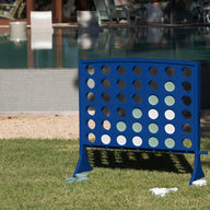 A large blue Ledge Lounger Four In A Row oversized outdoor game stands on the grass near a swimming pool, with white and green discs in play. This all-weather board features giant four-in-a-row action, while some discs and bags are scattered on the ground nearby.