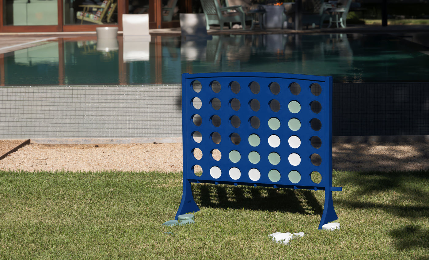 A large blue Ledge Lounger Four In A Row oversized outdoor game stands on the grass near a swimming pool, with white and green discs in play. This all-weather board features giant four-in-a-row action, while some discs and bags are scattered on the ground nearby.