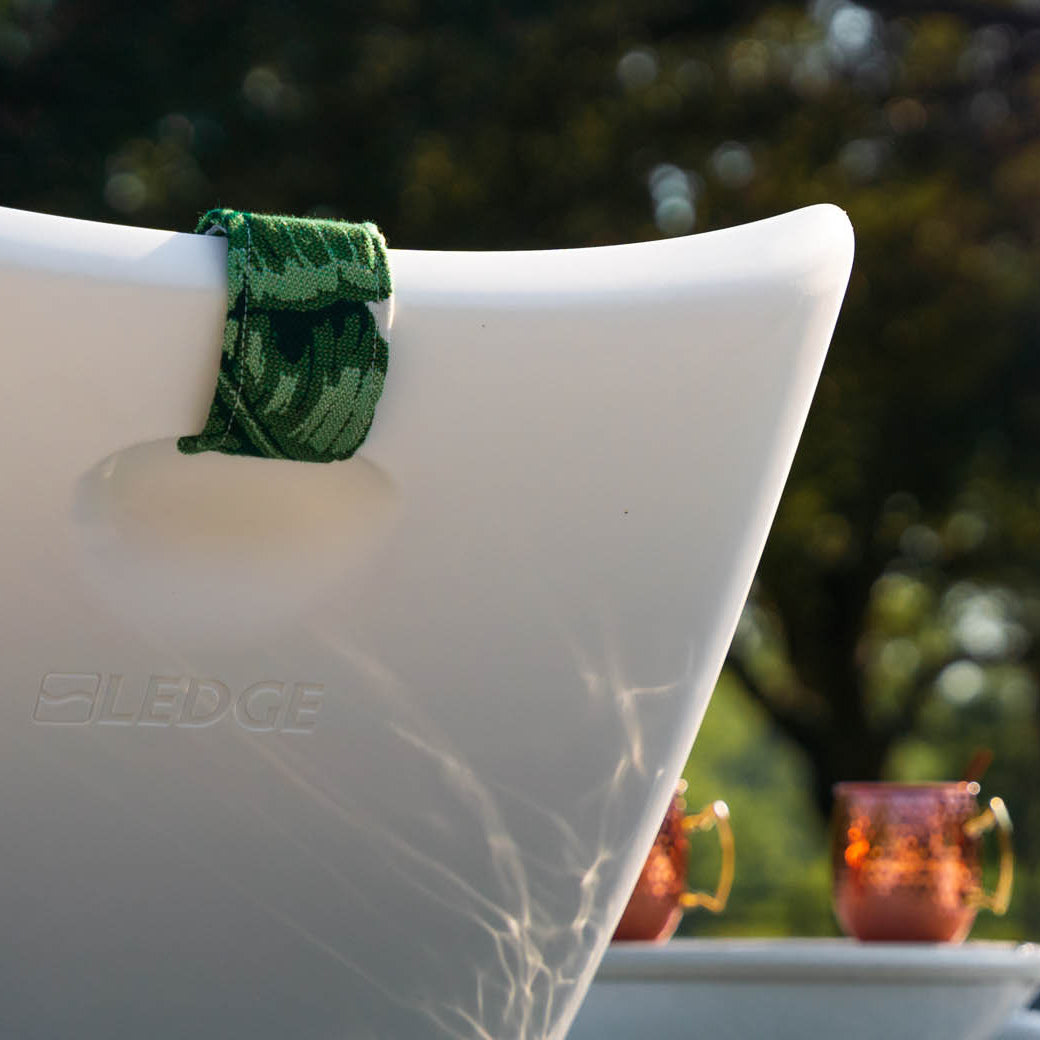 Close-up of a white Ledge Lounger chair with the Autograph Headrest Pillow, featuring a green towel on the backrest in an outdoor setting. Two copper mugs adorn a nearby table amidst sunlit greenery.
