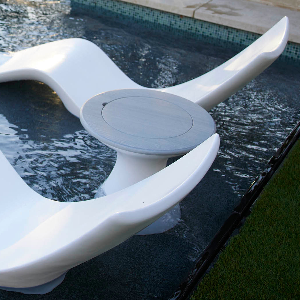 A sleek, white sculptural lounger set partially submerged in a shallow pool surrounds an Autograph Tall Ice Bin Side Table by Ledge Lounger. The pool is next to a grassy area and bordered by stone tiles, creating the perfect spot for relaxation and style.