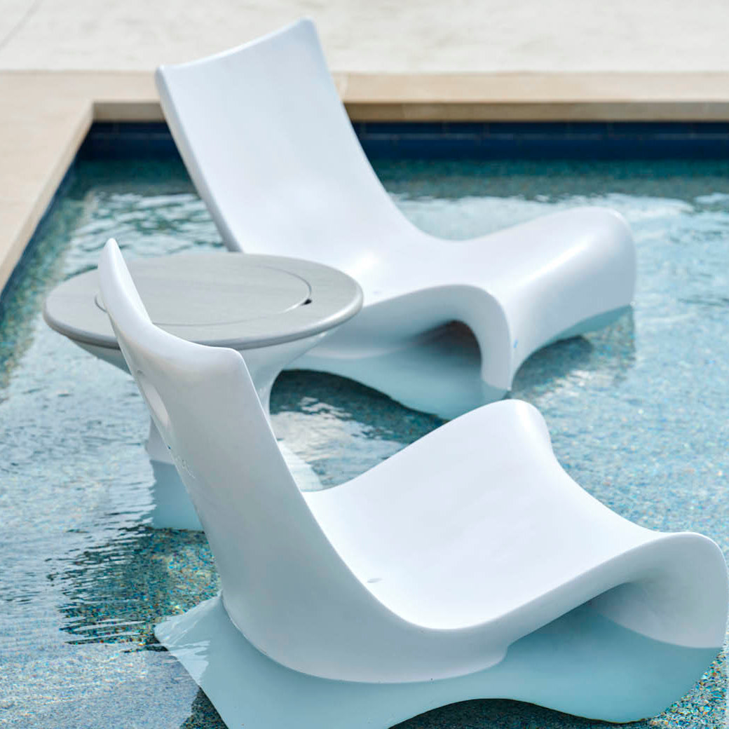 Two sleek Ledge Lounger white lounge chairs are partially submerged in a pool, flanking an Autograph Tall Ice Bin Side Table, creating a relaxing outdoor oasis.