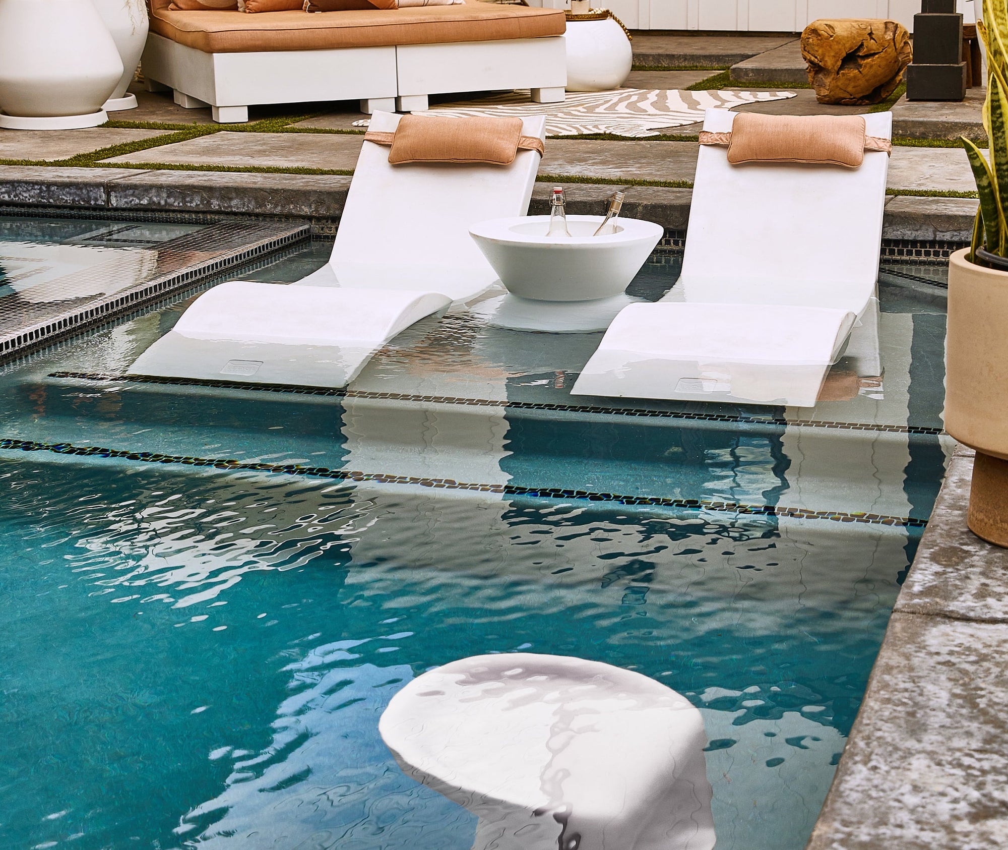 Two white lounge chairs with tan headrests are partially submerged in a pool, facing a white bowl with two bottles inside. Two underwater stools are visible in the foreground, and patio seating is in the background.