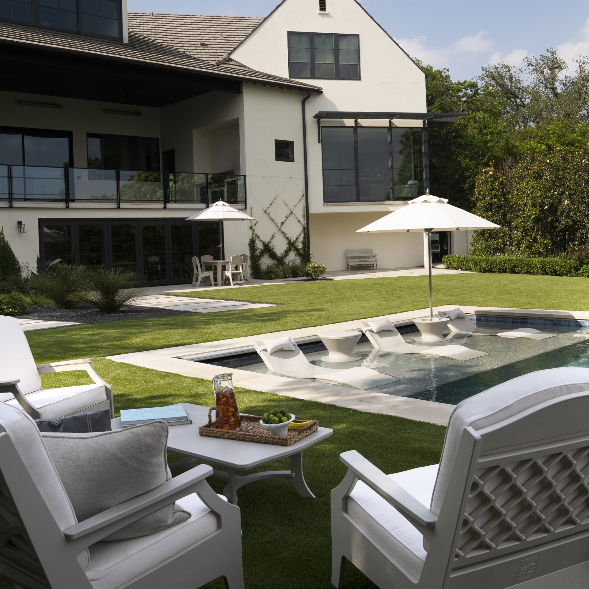A modern backyard with a swimming pool, white lounge chairs in and around the pool, side tables with drinks and snacks, umbrellas for shade, and a large house in the background.